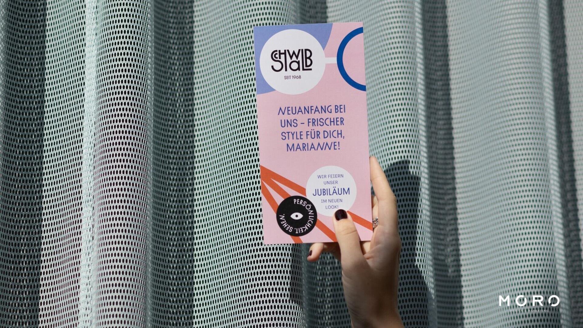 A hand with black nail polish holds a colorful flyer with geometric patterns and text in German, in front of a curtain with a mesh-like pattern. The flyer features bold fonts and modern design elements.