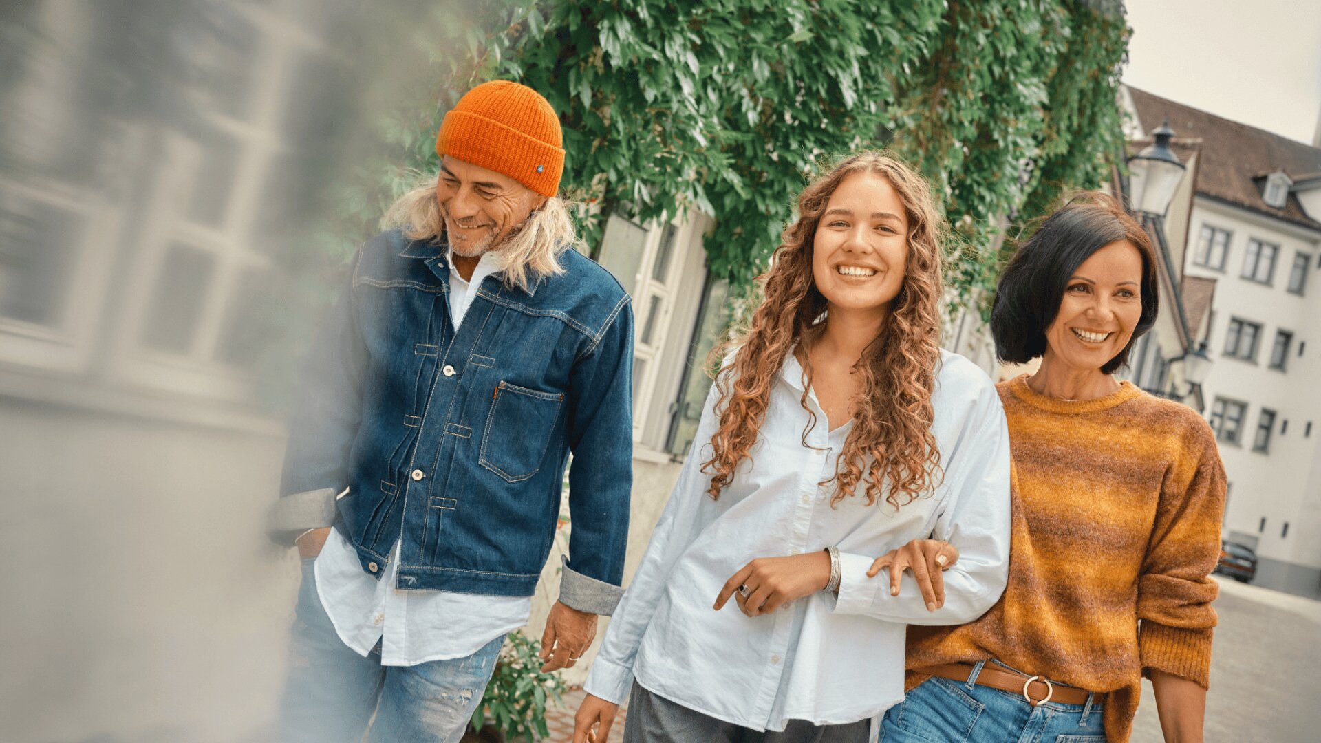 Drei Personen gehen im Freien spazieren, lächeln und verschränken die Arme. Zu der Gruppe gehören ein Mann in Jeansjacke und orangefarbener Mütze, eine junge Frau mit lockigem Haar und eine Frau in einem gestreiften Pullover. Im Hintergrund sind Bäume und Gebäude zu sehen.