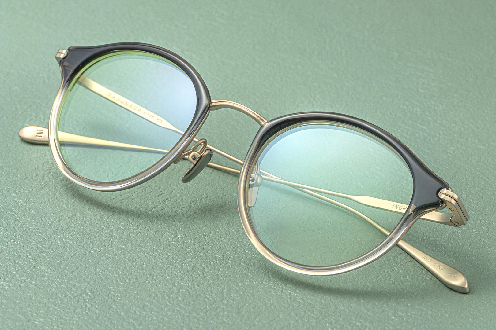 Die runde Brille mit dünnen goldenen Metallbügeln und durchscheinenden, blau getönten Gläsern ruht auf einer strukturierten grünen Oberfläche. Der Rahmen hat ein Farbverlaufsdesign, das oben schwarz und unten klar ist.