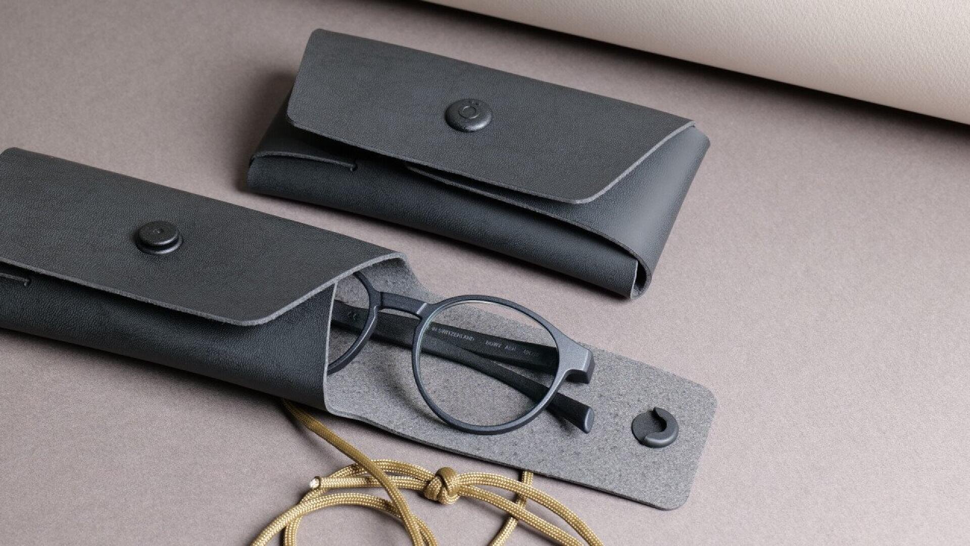 A pair of round black eyeglasses partially in a gray felt-lined black case, with another closed matching case nearby, all placed on a neutral-toned surface with a beige cord visible.