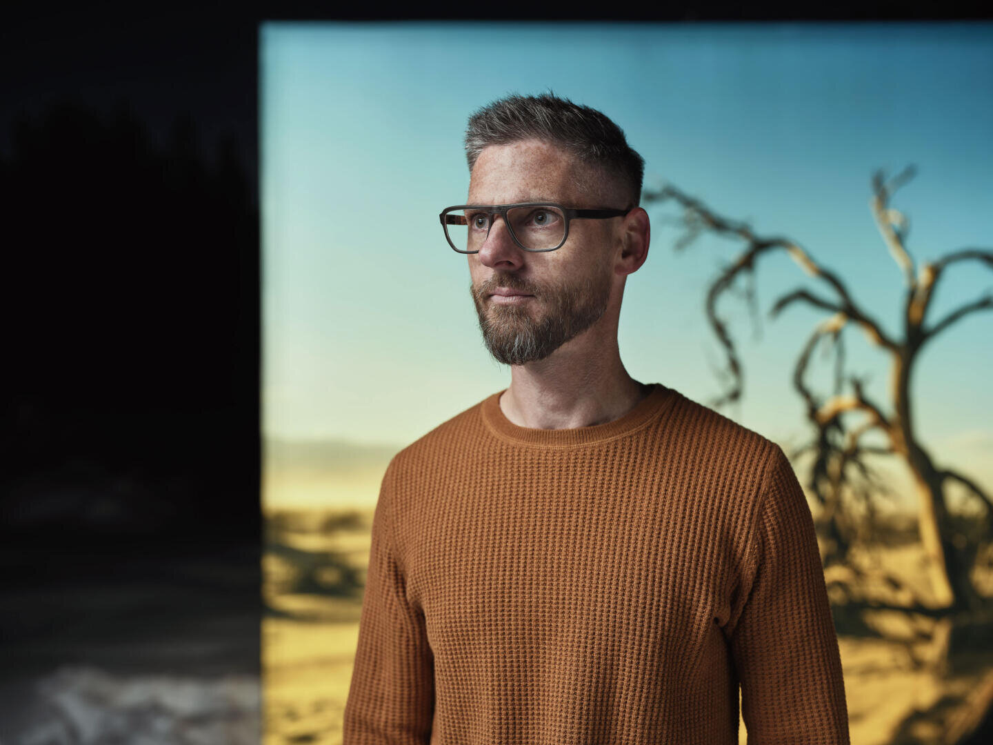 A man with short hair, glasses and beard, wearing a textured orange sweater, stands in front of a background showing a dry landscape with a leafless tree under a blue sky.
