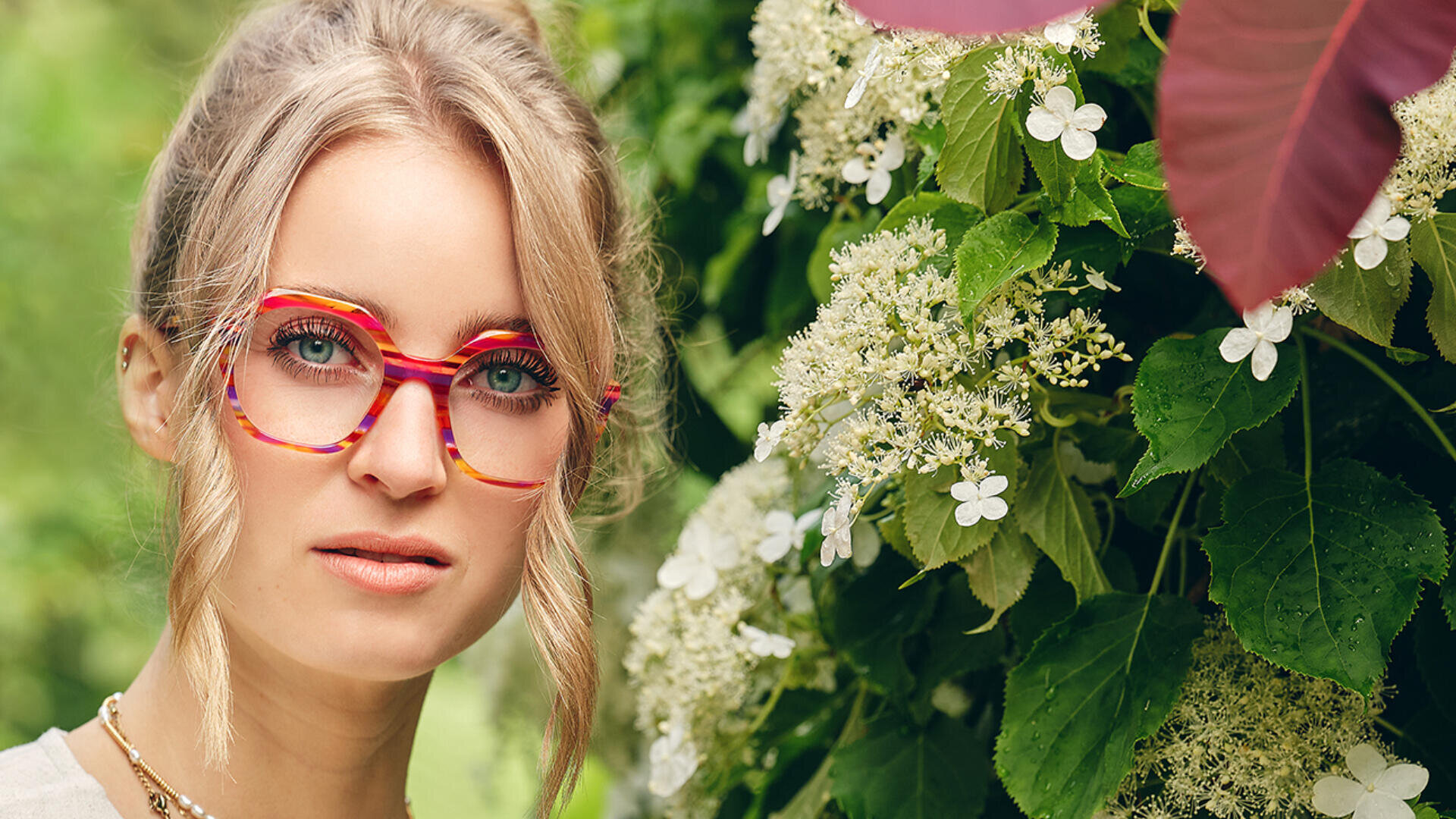 Eine Frau mit blondem Haar und bunter rot-gelber Brille steht neben üppigem grünem Laub und weißen Blumen und blickt leicht an der Kamera vorbei.