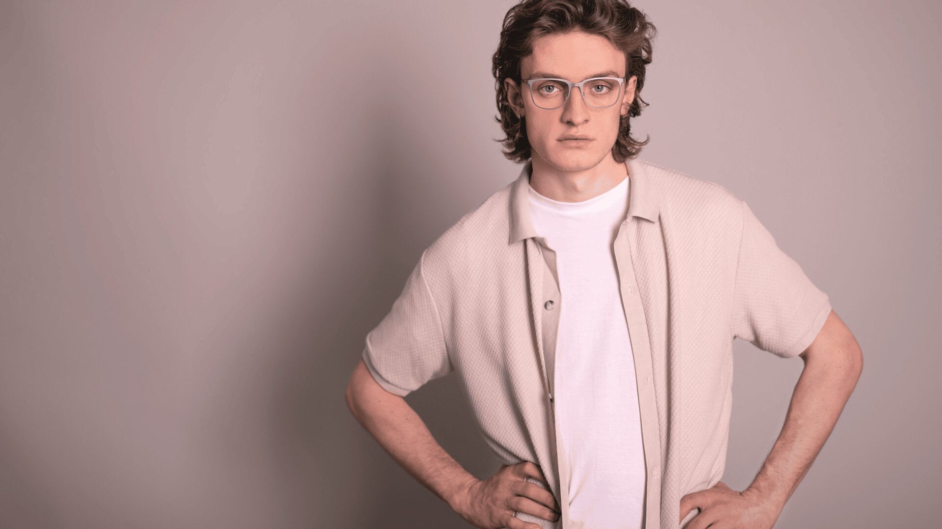 Eine junge Person mit gewelltem braunem Haar und Brille steht vor einem neutralen Hintergrund, trägt ein weißes T-Shirt unter einem hellen, kurzärmeligen Hemd, hat die Hände in die Hüften gestemmt und einen ernsten Gesichtsausdruck.