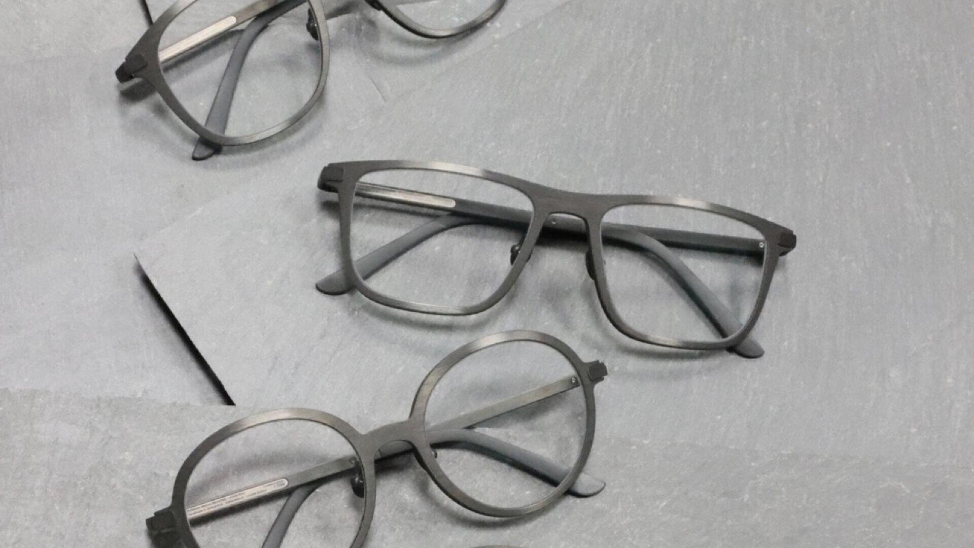 Three pairs of eyeglasses with black frames, each with different shapes (rectangular, round, and oval), are arranged on a flat, light gray surface.
