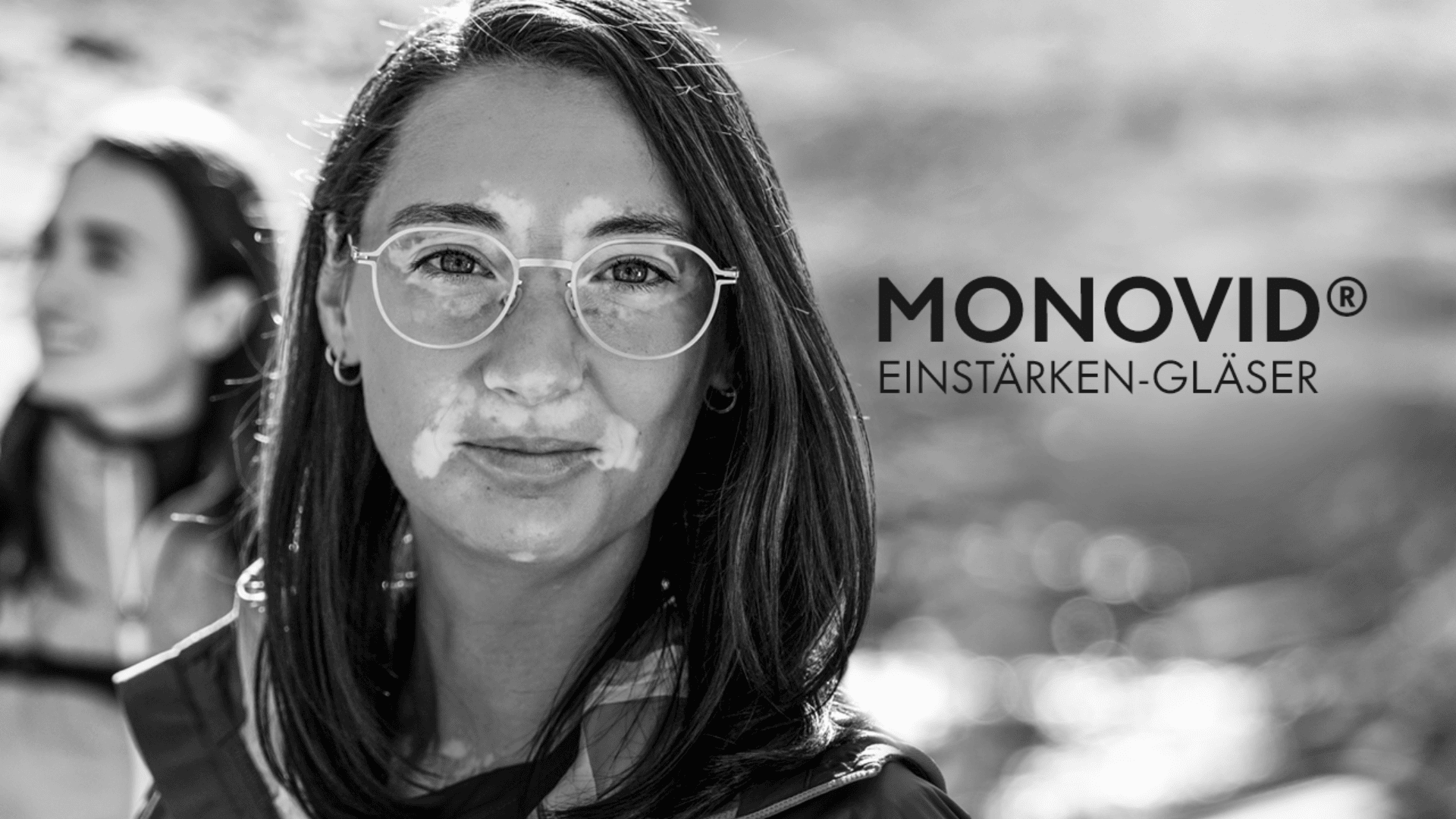 Schwarz-Weiß-Bild einer Frau mit Brille, die in die Kamera blickt. Im Hintergrund ist eine weitere Person unscharf zu sehen. Rechts neben ihrem Gesicht ist der Schriftzug MONOVID® EINSTÄRKEN-GLÄSER zu sehen.