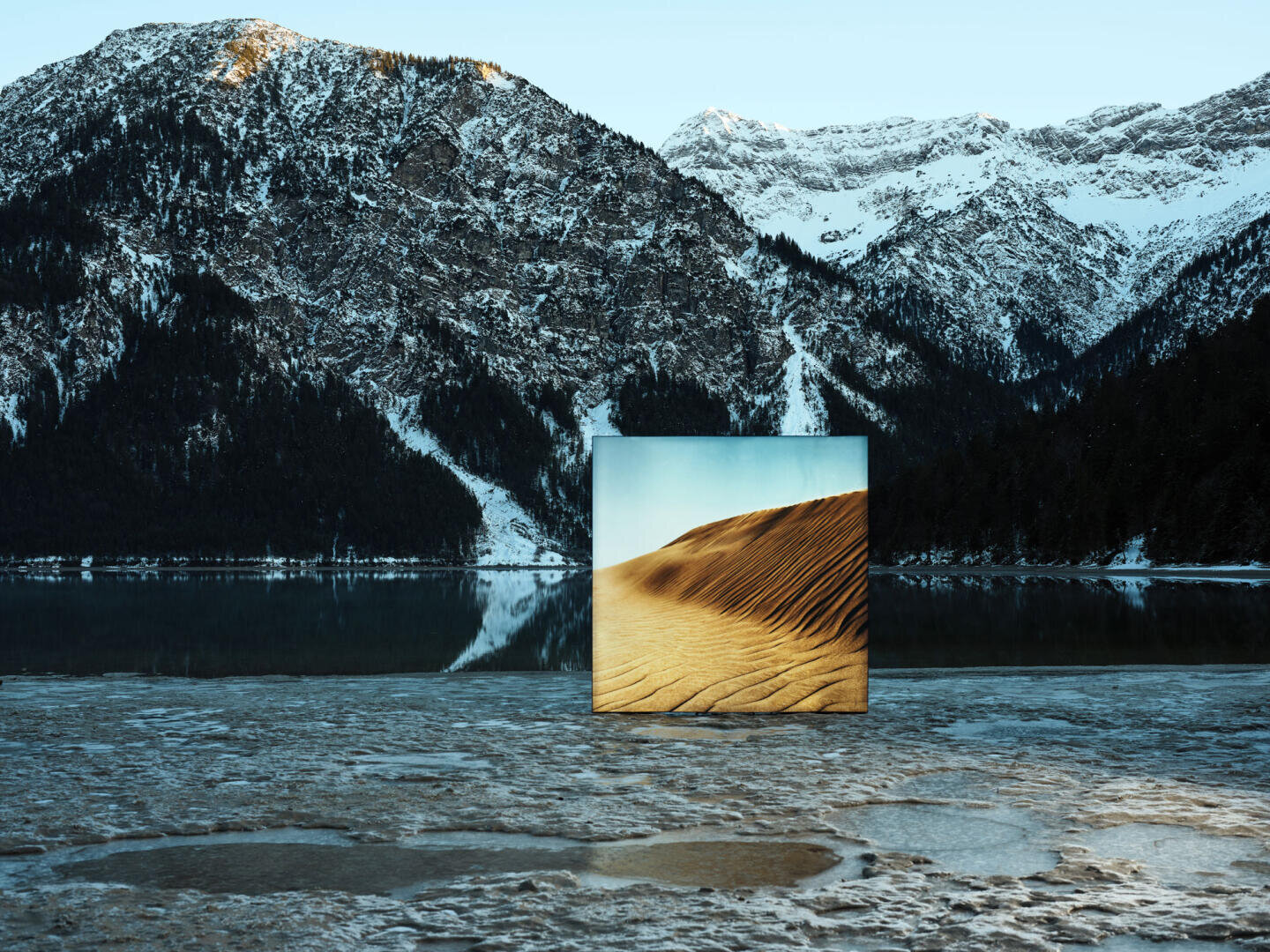 A square mirror stands on a snow-covered, frozen lake surrounded by mountains and reflects the image of a sunny desert sand dune, which stands in stark contrast to the wintry landscape.