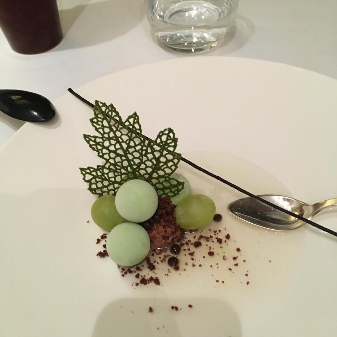 Ein plattiertes Dessert mit grünen Kugeln, grünen Trauben, Schokoladenstreuseln, einem dekorativen grünen Blatt und einem Stab aus dunkler Schokolade, serviert auf einem weißen Teller mit einem silbernen Löffel daneben.