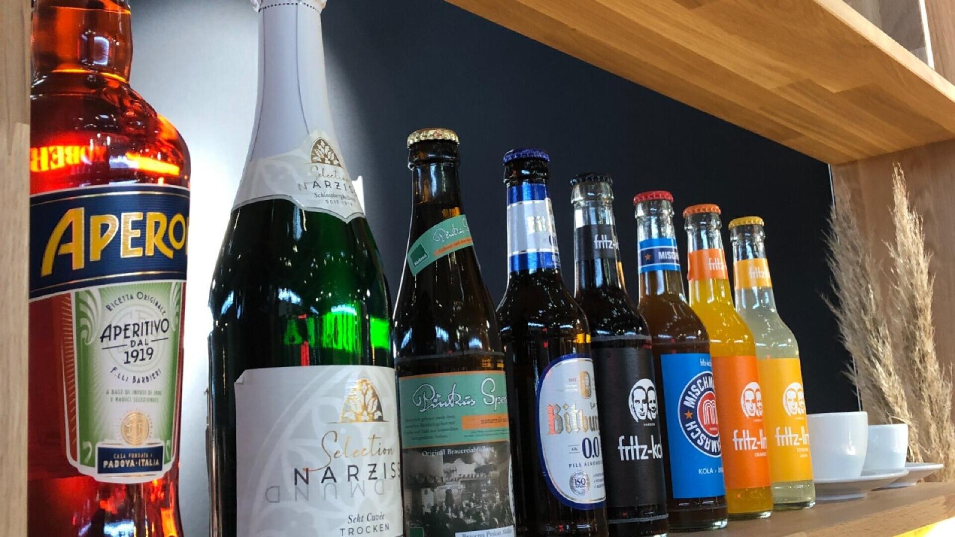 A row of assorted bottles, including Aperol, sparkling wine, several beers, and soft drinks, displayed on a wooden shelf with a dark background. Two white cups are visible at the end of the row.