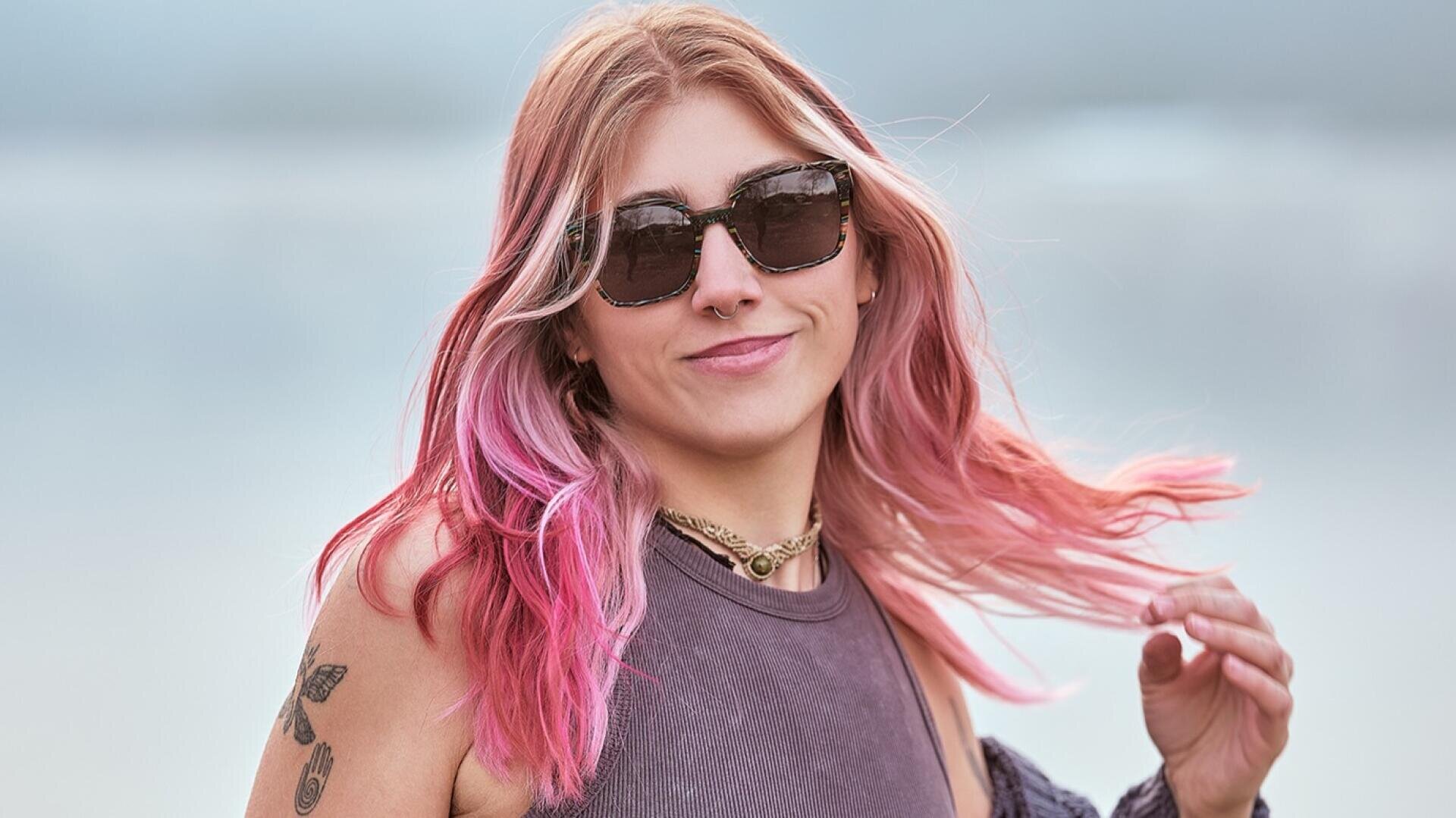 Eine junge Frau mit langen rosafarbenen Haaren, die eine Sonnenbrille und ein ärmelloses Oberteil trägt, lächelt in die Kamera. Sie hat Tätowierungen auf dem Arm, einen Nasenring und steht im Freien vor einem unscharfen Hintergrund.