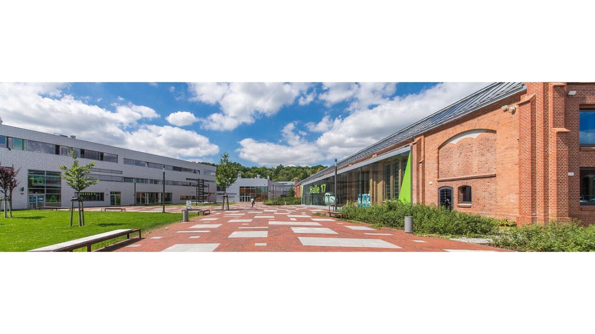 A spacious courtyard with red brick paving, modern white and glass buildings on the left, and a historic brick building on the right, under a blue sky with scattered clouds.