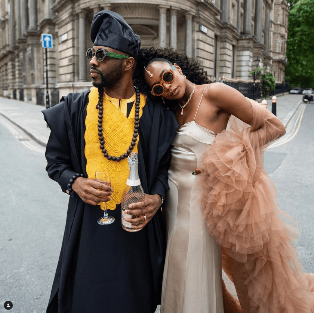 Ein stilvolles Paar posiert auf einer Straße in der Stadt; der Mann trägt traditionelle afrikanische Kleidung und eine Sonnenbrille und hält eine Flasche und ein Glas in der Hand, während sich die Frau in einem beigen Kleid an ihn lehnt und einen gerüschten Schal und eine große Sonnenbrille trägt.