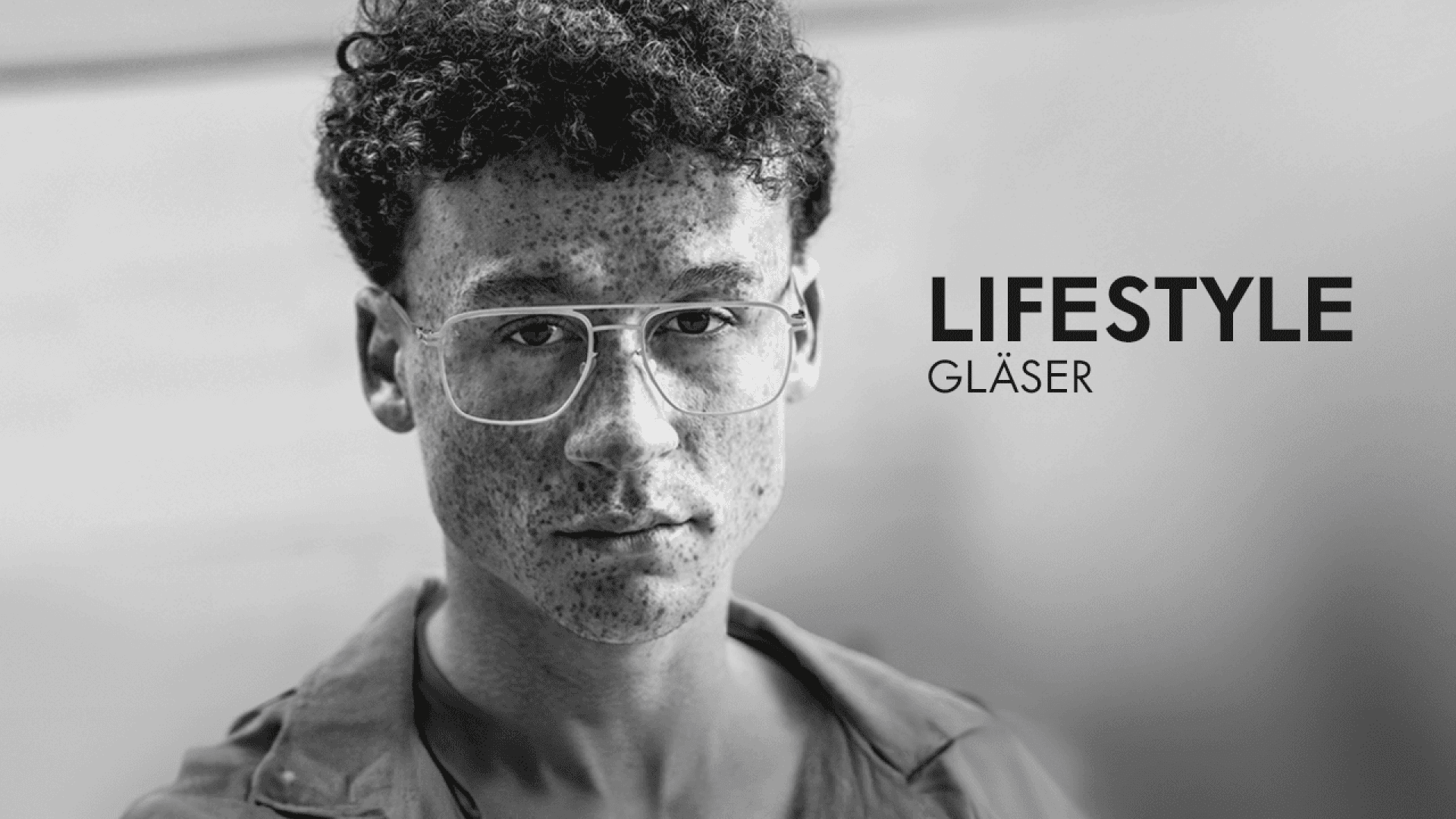 Black and white photo of a young man with short curly hair, freckles, and clear eyeglasses, looking directly at the camera. The words LIFESTYLE GLÄSER appear beside him.