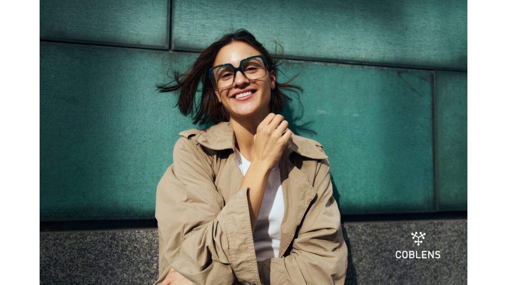 A smiling woman wearing large black glasses and a beige coat stands against a teal wall. Her hair is slightly windblown, and sunlight shines on her face. The COBLENS logo appears in the bottom right corner.