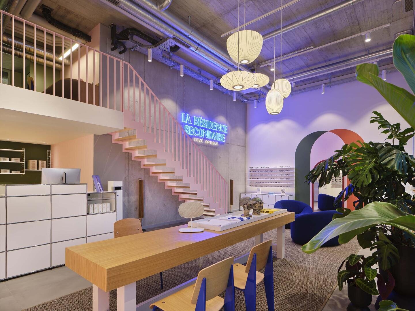 Modernes Büro mit Holztisch, blauen Stühlen, großer Pflanze, Pendelleuchten, pastellfarbener Treppe und Leuchtreklame mit der Aufschrift "LA RÉSIDENCE SHOW PLACE". Der Raum hat freiliegende Decken und ein farbenfrohes, modernes Dekor.