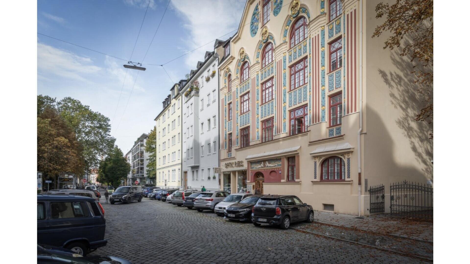 A cobblestone street lined with parked cars runs beside colorful, ornate buildings, including one with intricate patterns and arched windows, under a partly cloudy sky with trees on the left side.