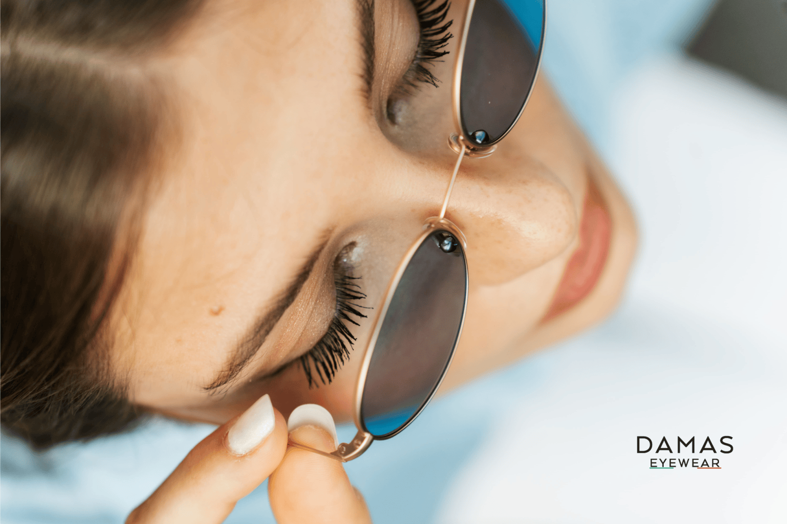 Nahaufnahme einer Frau, die eine Sonnenbrille mit goldenen Rändern trägt und diese mit der Hand zurechtrückt. Ihre langen Wimpern und ihre helle Haut sind zu sehen. Unten rechts erscheint das Logo von DAMAS EYEWEAR.