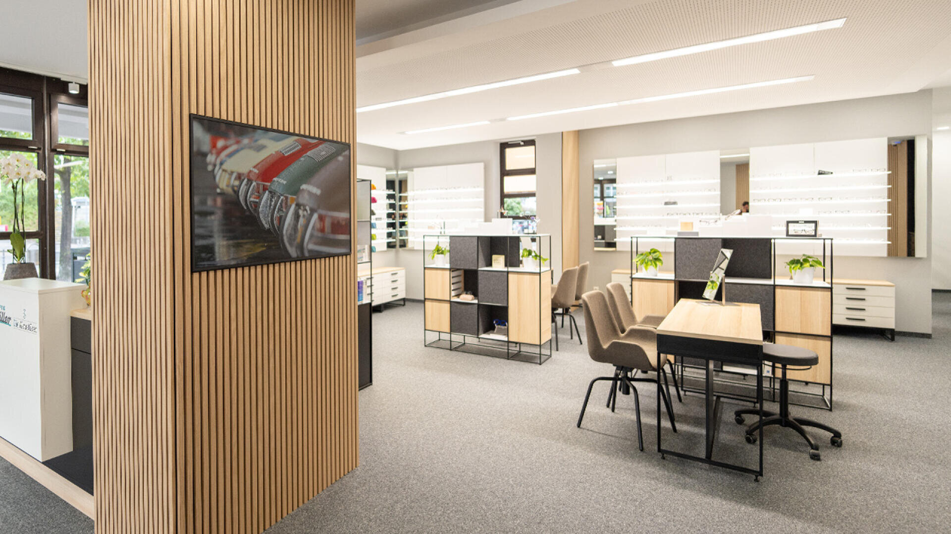 Eine moderne, gut beleuchtete Optikerpraxis mit Brillenregalen, Holzakzenten, einem Konferenztisch mit Stühlen, Topfpflanzen und einem Fernseher an der Wand. Große Fenster sorgen für Tageslicht.