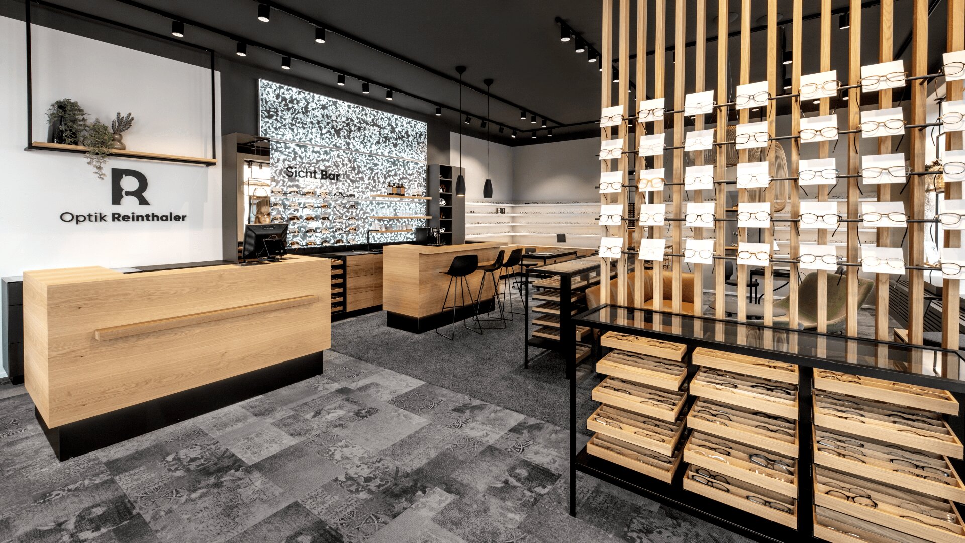 Ein modernes Optikergeschäft mit Holztheken, Brillenregalen, Sitzbereich und einem großen beleuchteten Bildschirm im hinteren Bereich. Die Einrichtung ist minimalistisch in Schwarz und Holz gehalten.