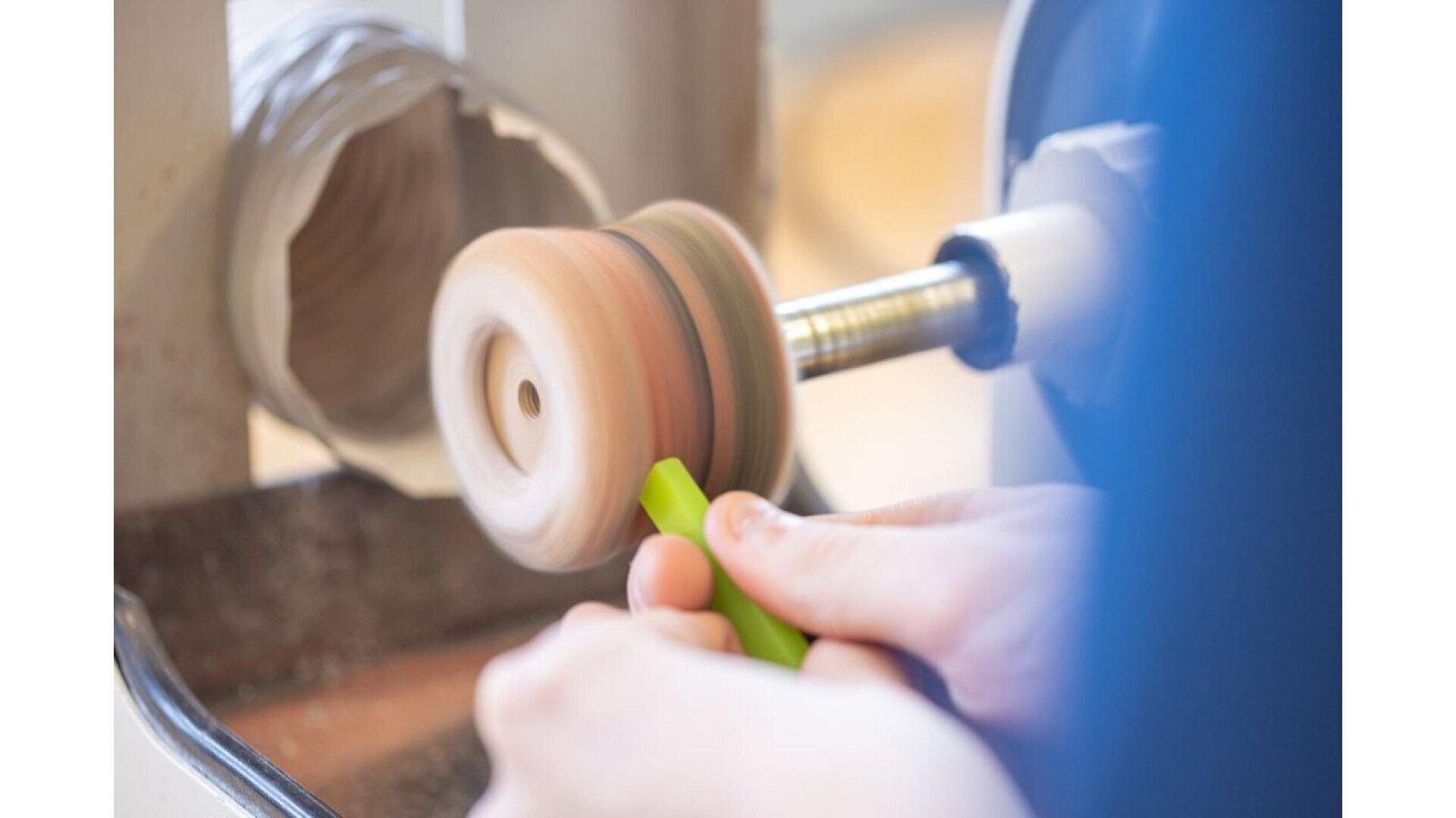 Close-up of hands polishing a spinning wooden object on a lathe, holding a green tool against the wood as it rotates, with wood shavings visible below.