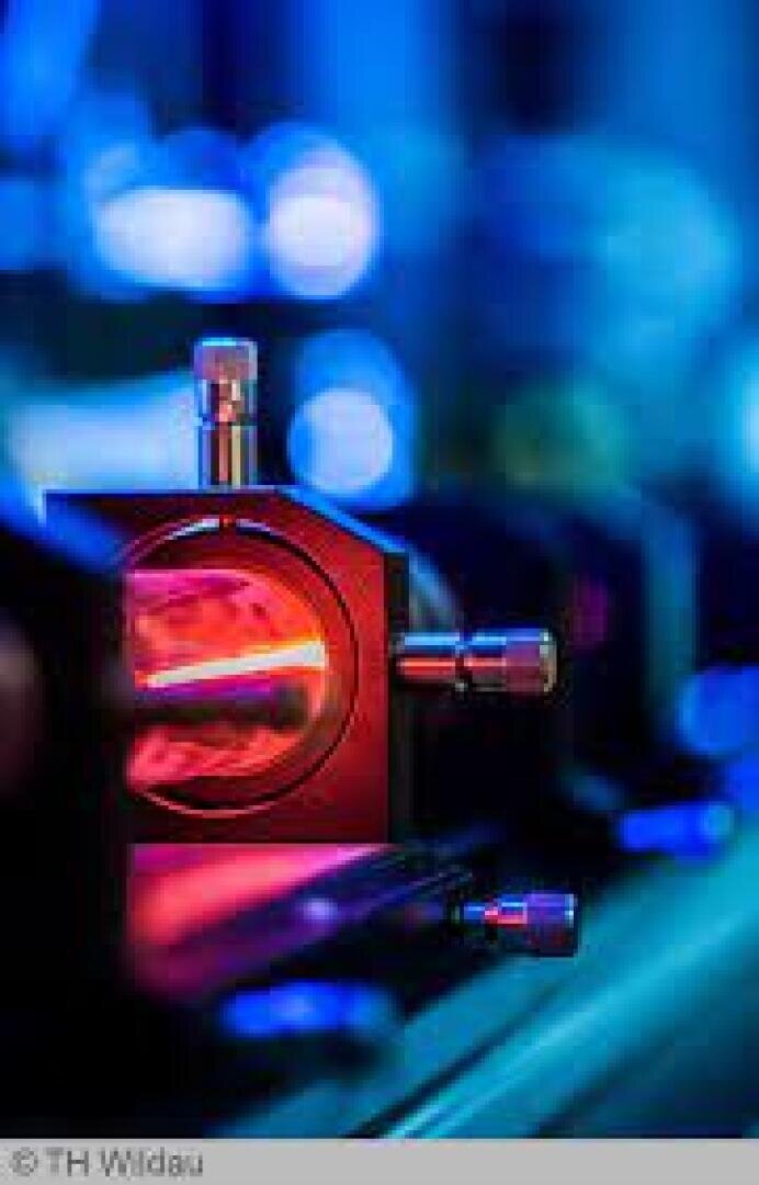 Nahaufnahme einer wissenschaftlichen Ausrüstung mit einem glühenden roten Laserstrahl, der durch eine Metall- und Glasapparatur verläuft, vor einem unscharfen blauen Hintergrund.