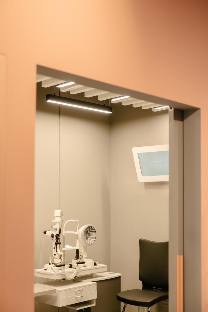 Ein modernes Untersuchungszimmer eines Optometristen mit einem Sehtestgerät auf einem Schreibtisch, einem schwarzen Stuhl und einer beleuchteten Sehtafel an der Wand. Der Raum hat neutral gefärbte Wände und eine moderne Beleuchtung.