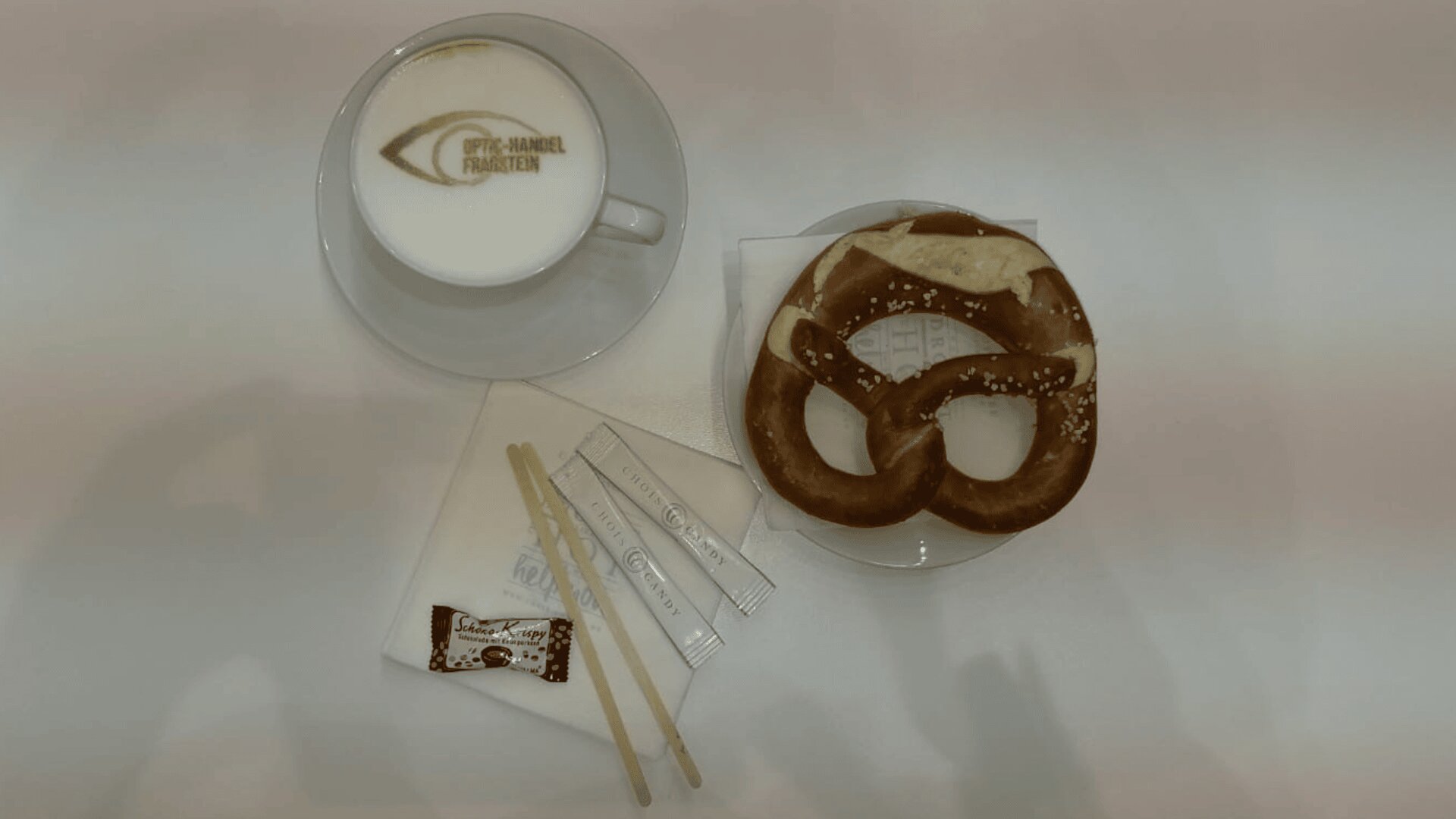 Eine Tasse Cappuccino mit Latte Art und OPHTHALMOL FRANKFURT Schriftzug, eine weiche Brezel auf einer Serviette, Zuckerpäckchen, ein Schokoladenquadrat und zwei Kaffeerührer auf einem weißen Tisch.