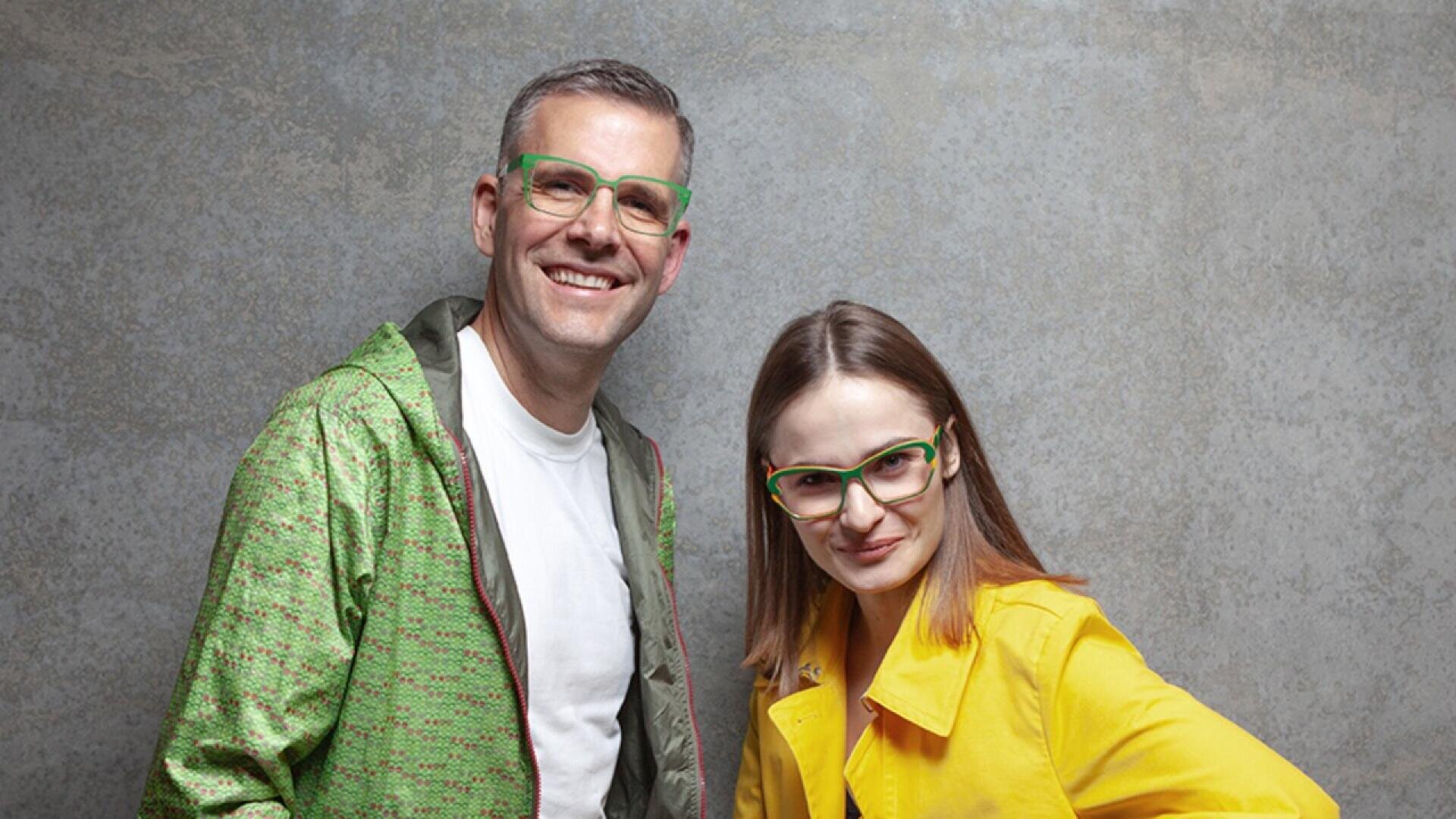 A smiling man in a green jacket and glasses stands next to a woman in a yellow coat and green glasses, both posing in front of a gray textured background.