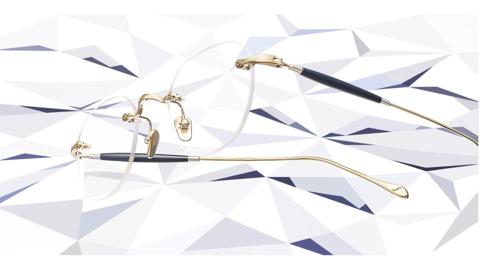 A pair of rimless eyeglasses with gold temples and black arms displayed against a white, faceted geometric background.
