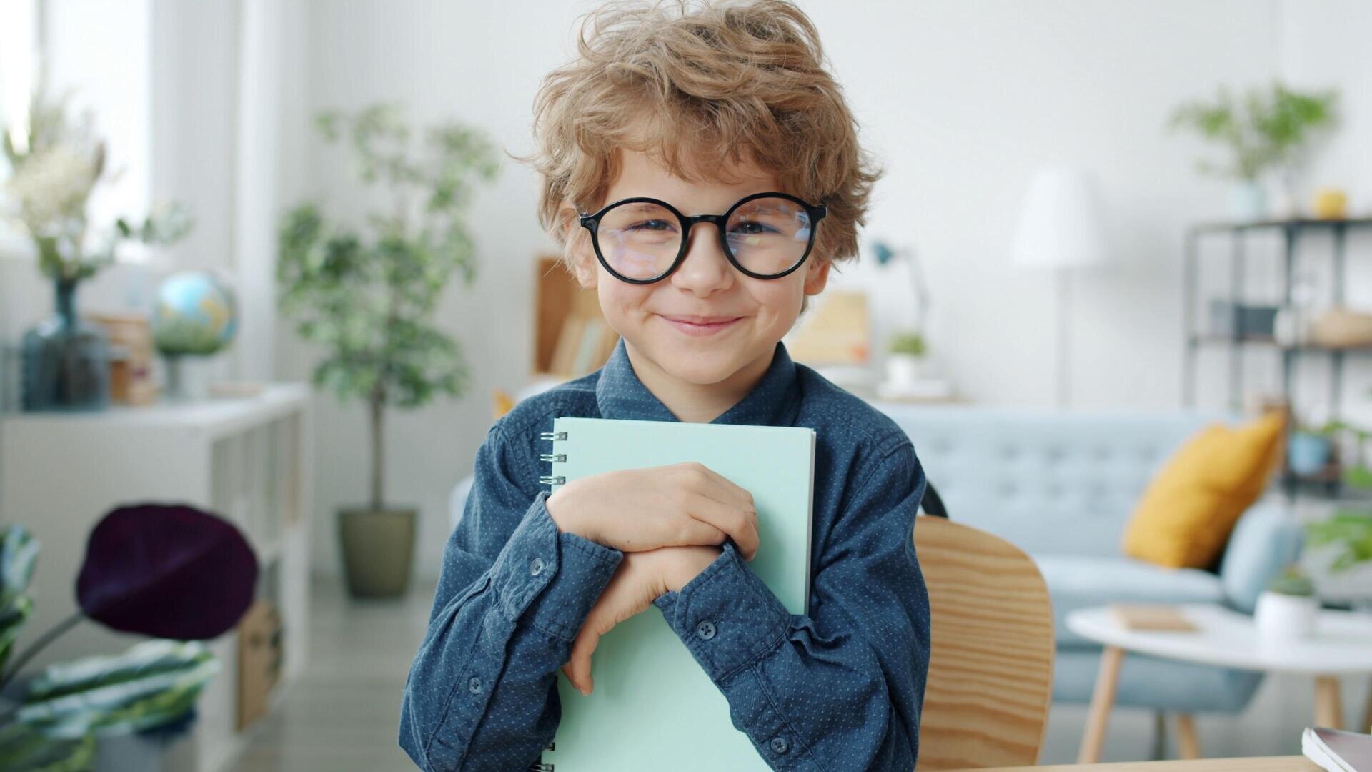 Ein lächelnder kleiner Junge mit lockigem Haar und großer runder Brille umarmt ein hellblaues Notizbuch, während er an einem Schreibtisch in einem hellen, gemütlichen Raum mit Pflanzen und modernem Dekor sitzt.