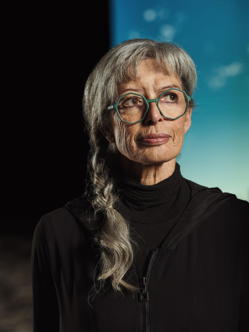 An older woman with long gray hair braided into a pigtail wears blue-green glasses and a black top and looks thoughtfully to the side. The background is half dark and half blue with a soft, blurred effect.
