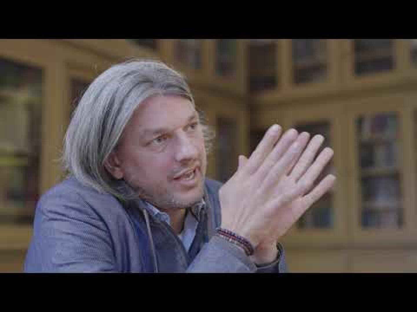 Ein Mann mit schulterlangen grauen Haaren und kurzem Bart, der eine blaue Jacke trägt, sitzt in einem Innenraum vor einem Bücherregal und gestikuliert mit seinen Händen, während er spricht.