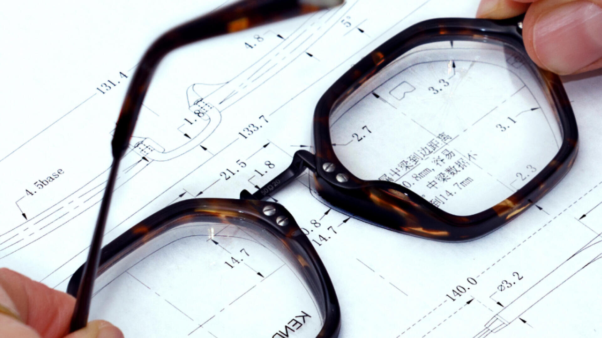 A close-up of hands holding eyeglass frames over a technical drawing with measurements and diagrams, suggesting eyewear design or engineering.