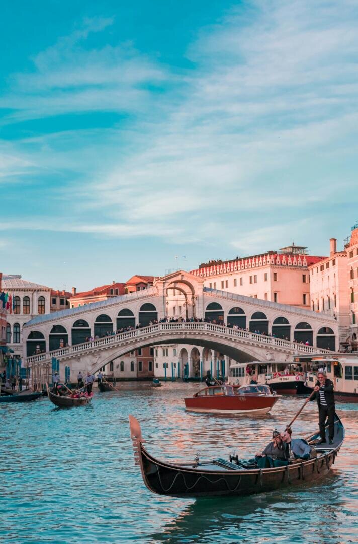 Ein Gondoliere steuert eine Gondel mit Passagieren durch einen Kanal in Venedig, mit der historischen Rialtobrücke und den umliegenden Gebäuden unter blauem Himmel im Hintergrund.
