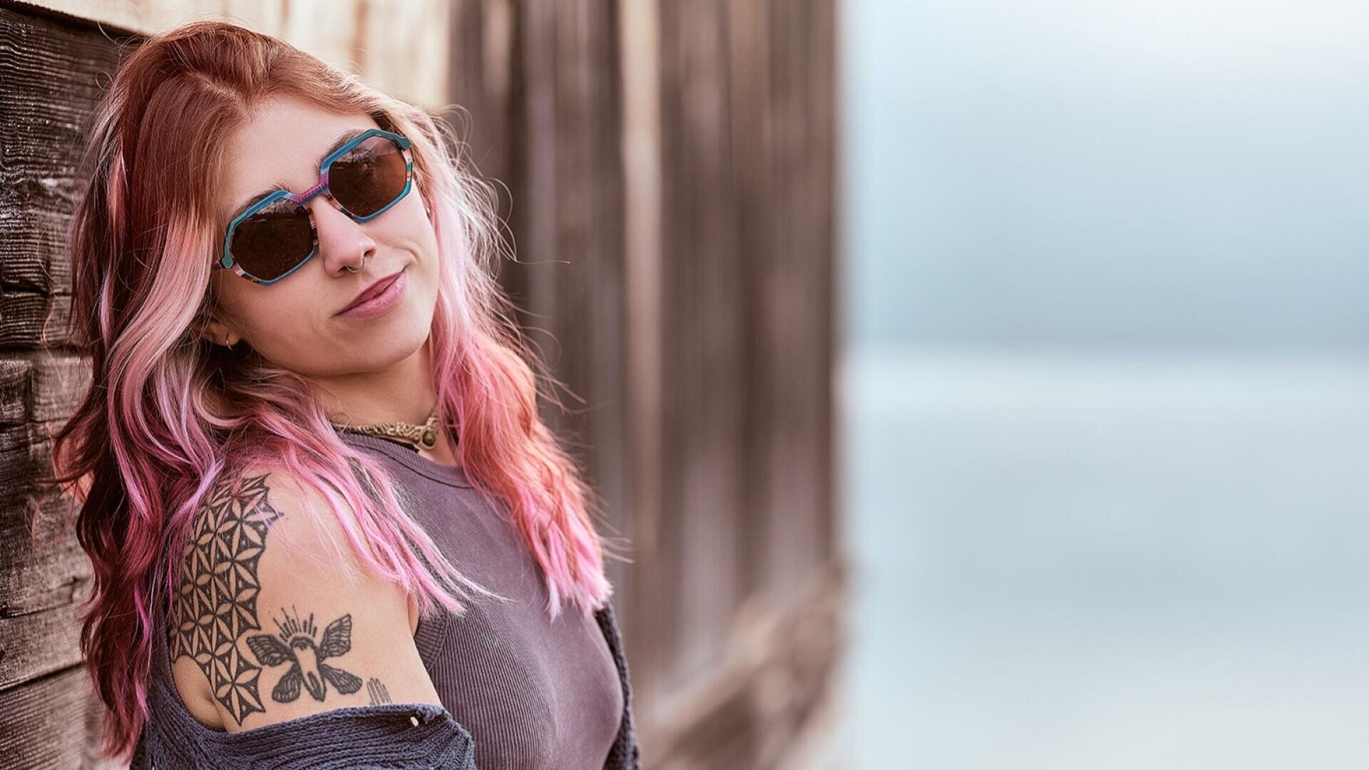 Eine Frau mit rosa und blondem Haar, die eine Sonnenbrille und ein ärmelloses Oberteil trägt, lehnt an einer Holzwand. Sie hat eine geometrische und eine Schmetterlings-Tätowierung auf ihrem Oberarm. Der Hintergrund ist unscharf und hat einen ruhigen, hellblauen Ton.