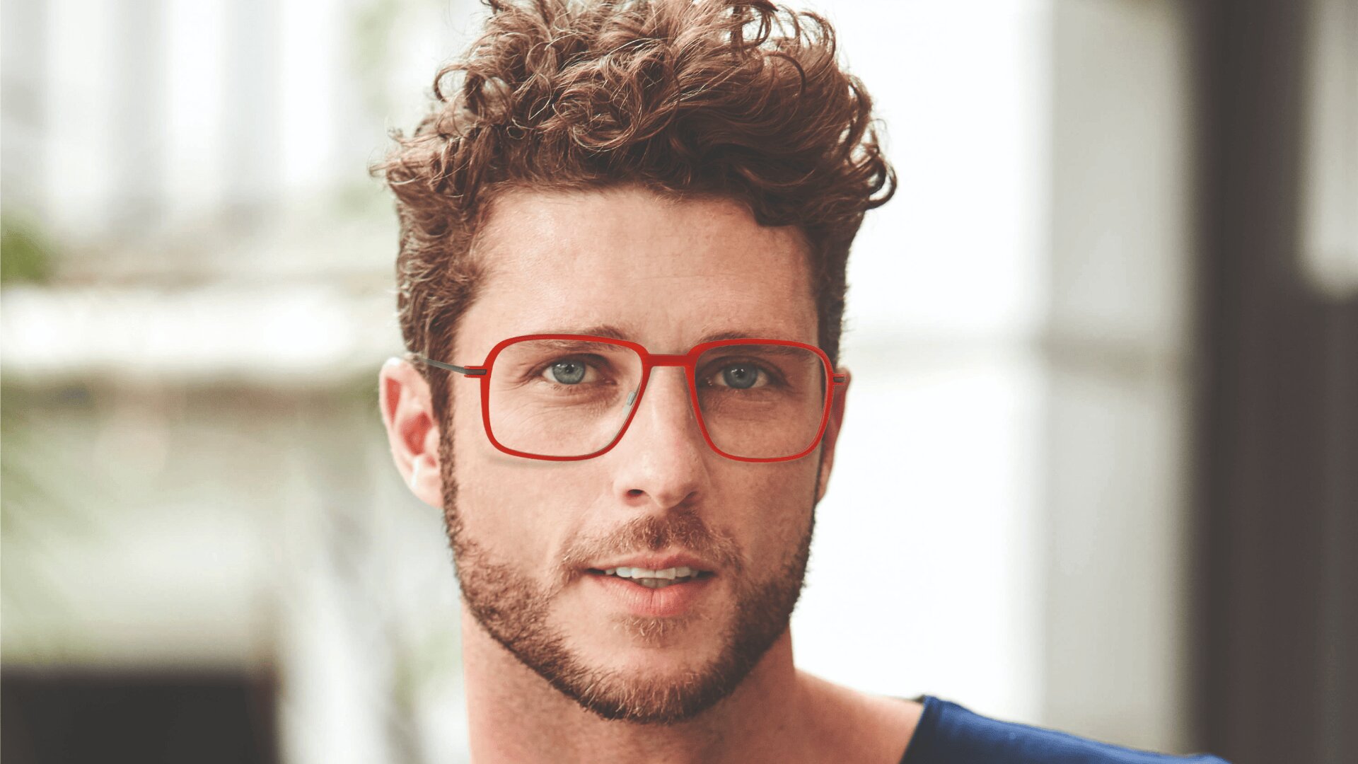 Ein Mann mit lockigen braunen Haaren, einem kurzen Bart und einer rotgeränderten Brille blickt mit neutralem Blick in die Kamera. Der Hintergrund ist unscharf und in hellen Farben gehalten.