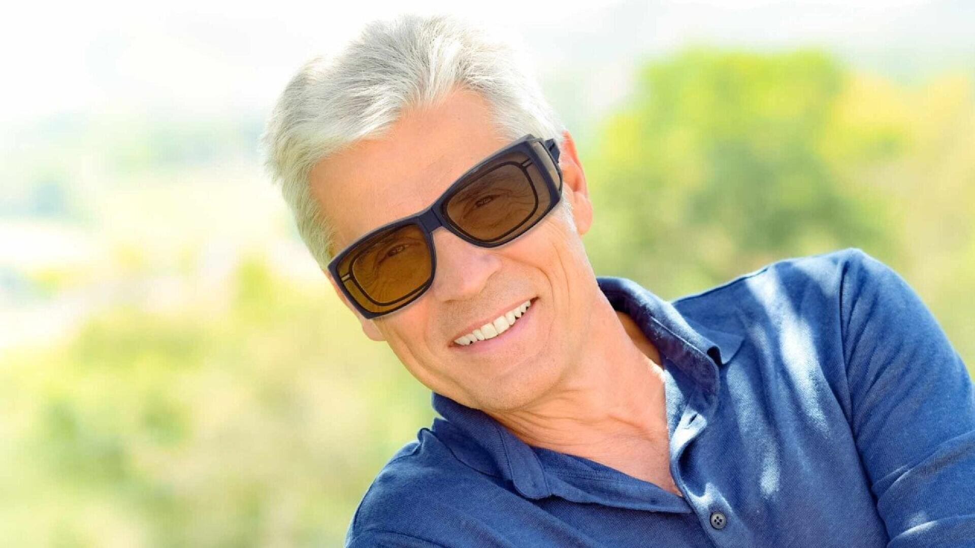 Smiling older man with short gray hair wearing sunglasses and a blue collared shirt, outdoors with a blurred green and yellow background.