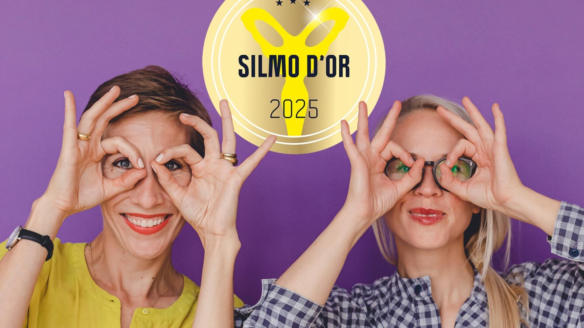 Zwei Frauen stehen vor einem lilafarbenen Hintergrund und formen mit ihren Händen über den Augen eine Brille. Über ihnen ist ein gelbes SILMO DOR 2025-Logo zu sehen. Beide lächeln und wirken fröhlich.