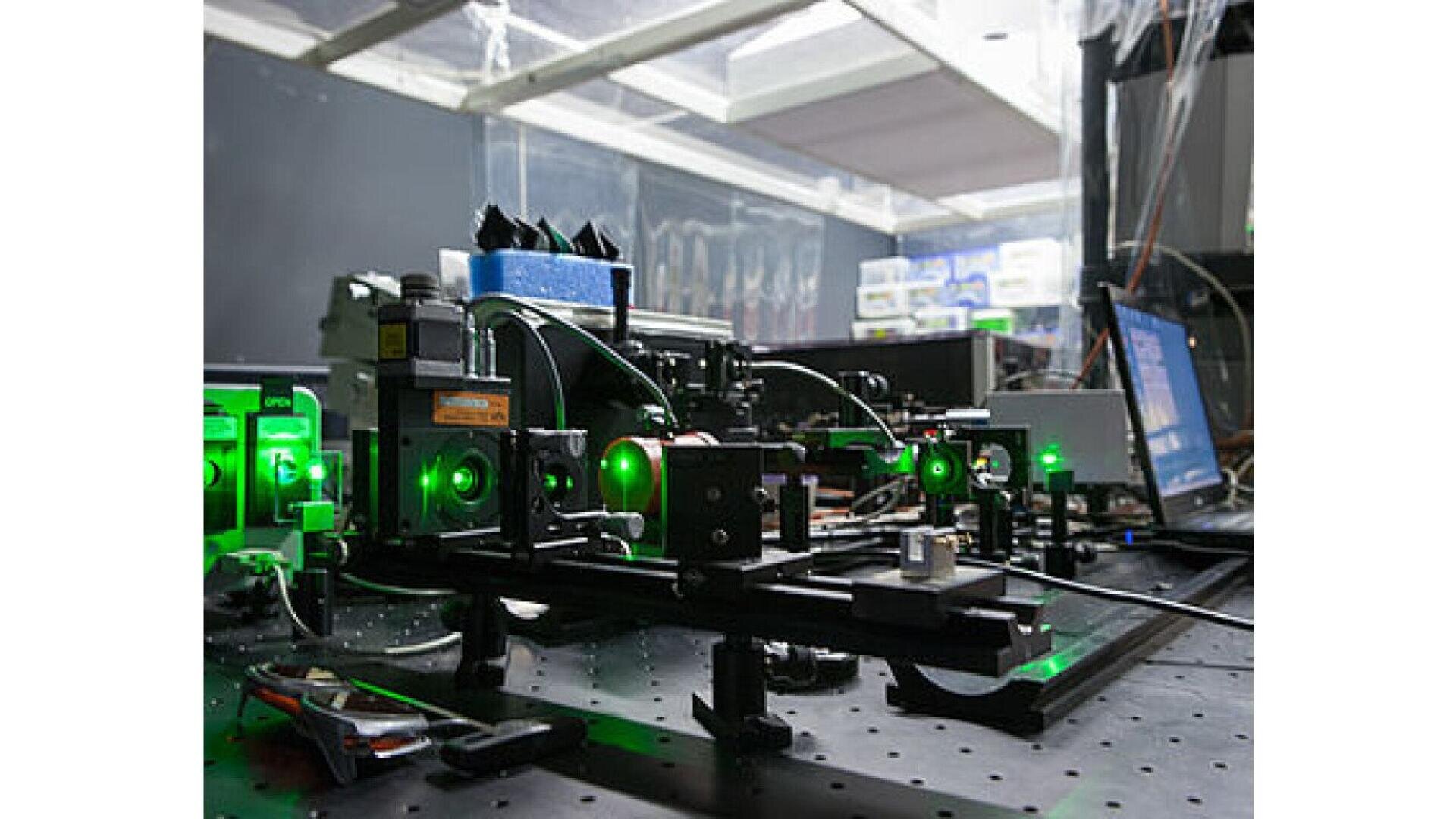 A laser optics laboratory setup with green lasers shining through various lenses, mirrors, and equipment on a black perforated table, surrounded by wires and electronic devices.