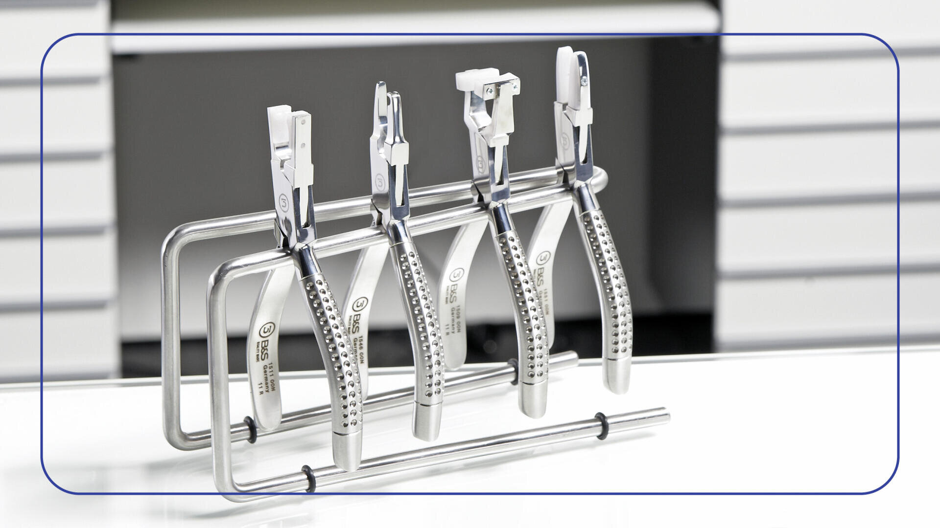 Vier Metallzahnpinzetten sind ordentlich auf einem Metallständer angeordnet, der auf einer weißen Fläche mit einem Hintergrund aus weißen Schubladen steht. Die Pinzetten haben strukturierte Griffe und sind aufrecht in einer Reihe angeordnet.