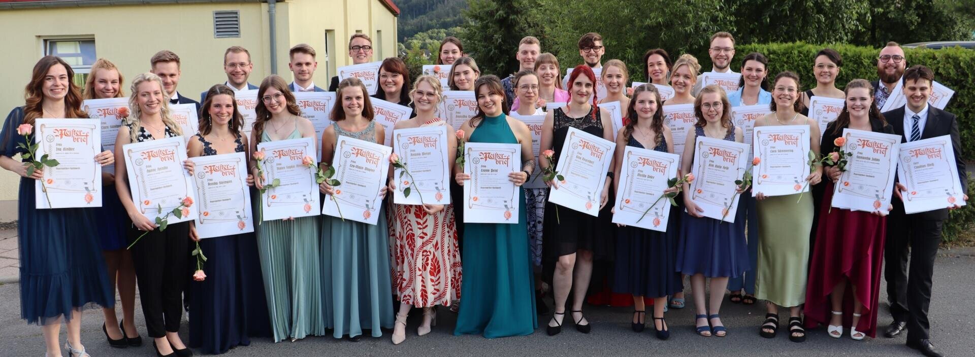 Eine Gruppe junger Erwachsener in formeller Kleidung steht im Freien, hält große Zertifikate und einzelne Rosen in den Händen und lächelt in die Kamera. Im Hintergrund sind Bäume und ein gelbes Gebäude zu sehen.