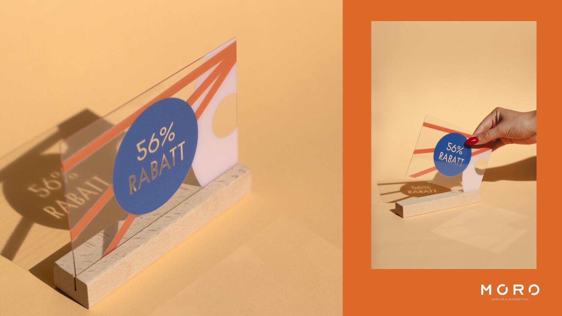 A clear sign with 56% RABATT in white on a blue circle, featuring abstract orange and yellow shapes, stands in a wooden base on a beige background. On the right, a hand holds the sign. MORO logo appears at the bottom right.