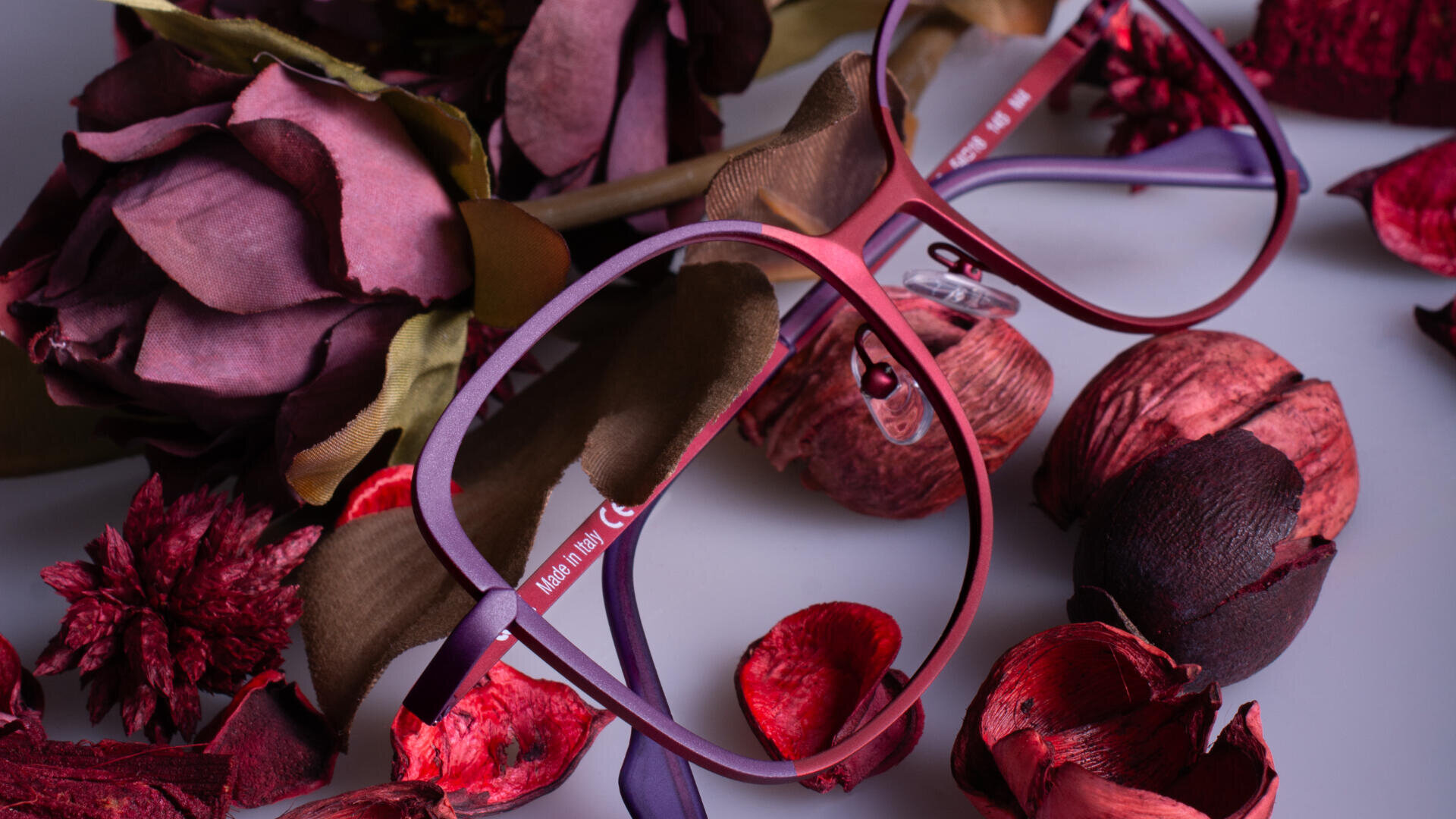 A pair of red-framed eyeglasses is placed among dried, burgundy and red flowers and petals on a light surface. The scene has a warm, artistic, and autumnal feel.