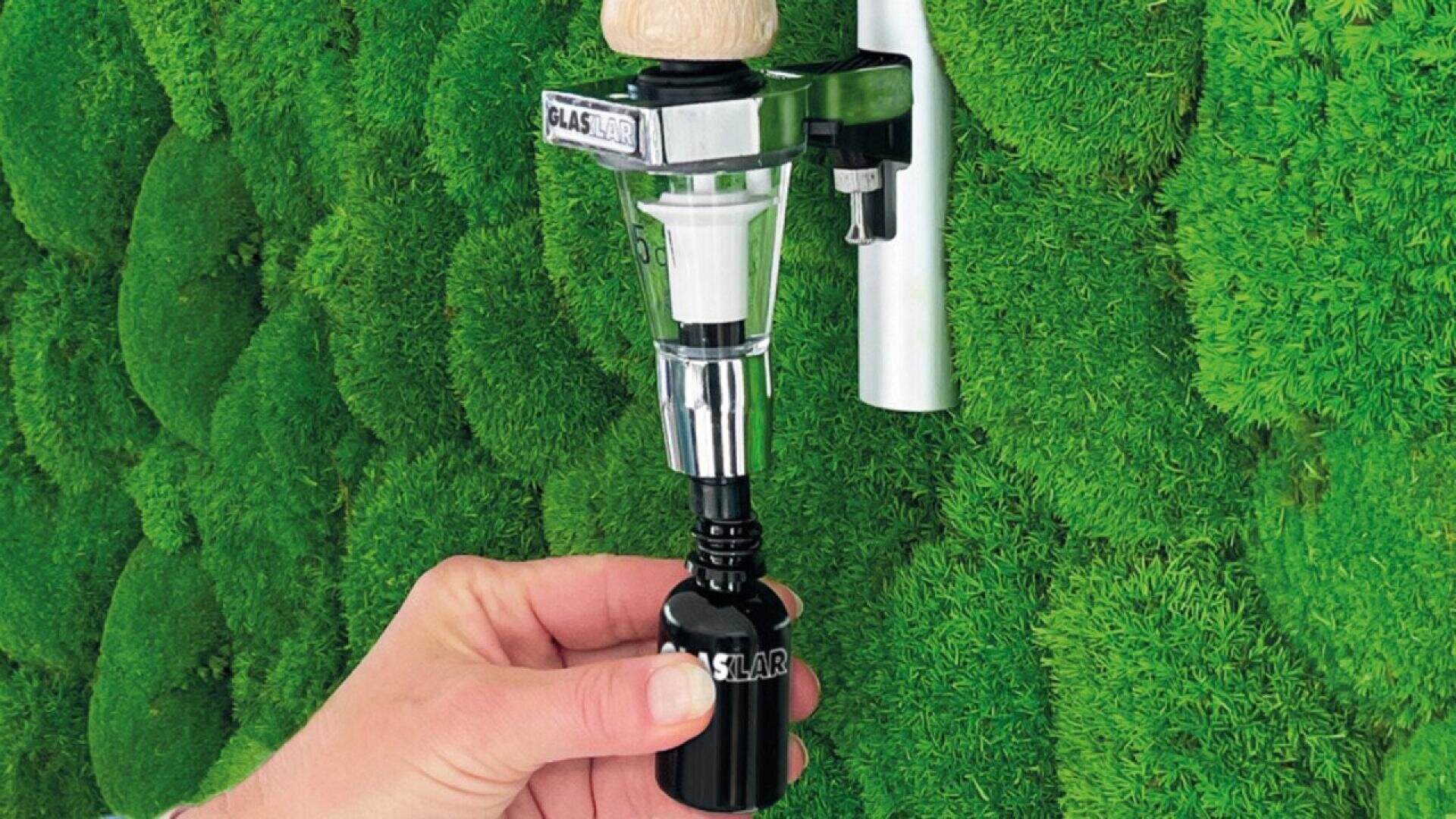 A hand fills a small black glass bottle using a wall-mounted dispenser attached to a green, moss-covered wall.