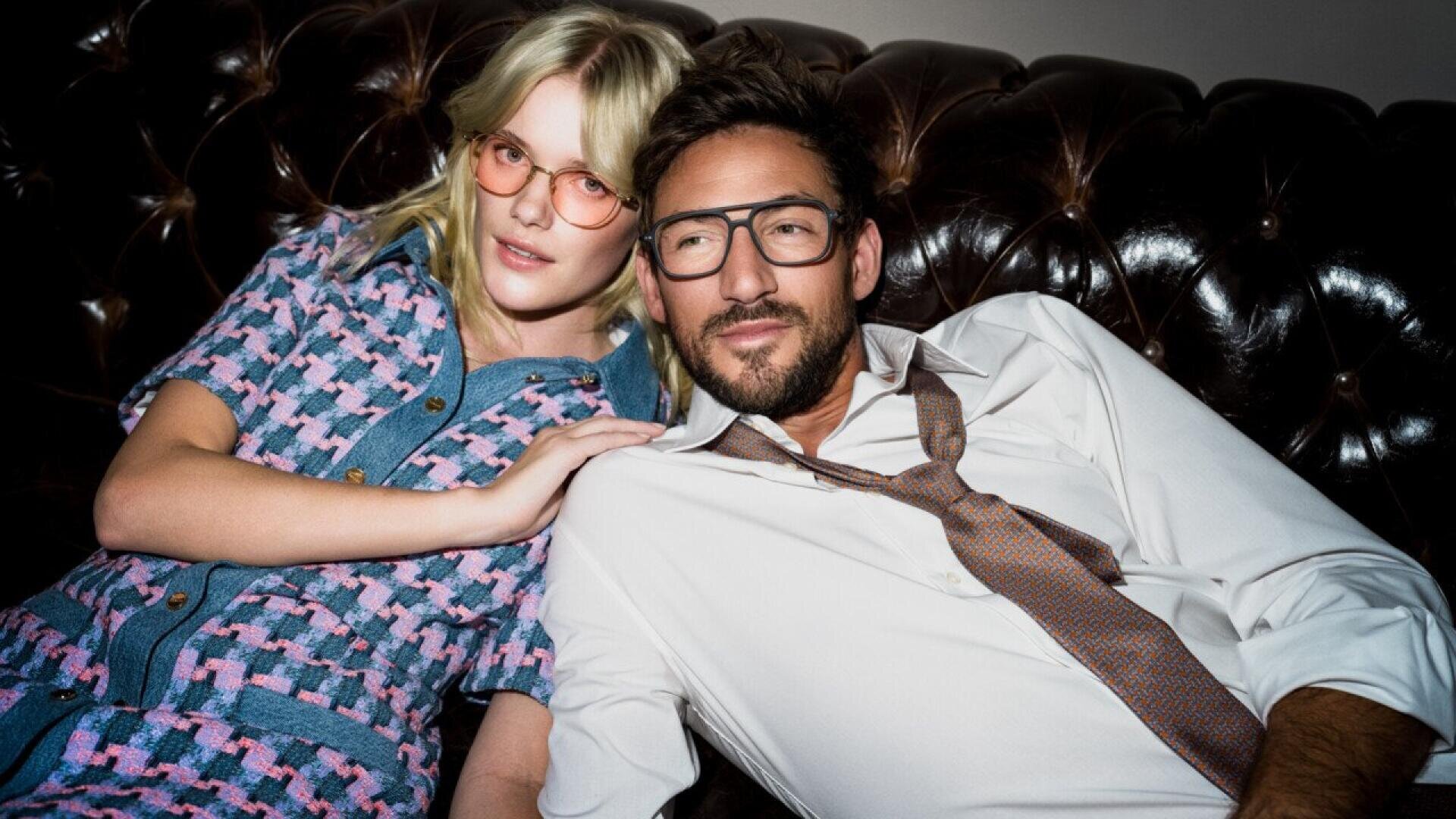A woman in a pink and blue patterned dress and a man in a white shirt and loosened tie, both wearing glasses, relax on a dark leather sofa, leaning close together and looking off to the side.