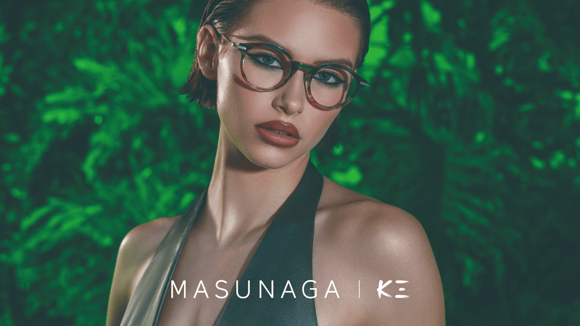 Eine Frau mit kurzen braunen Haaren und auffälligem Make-up trägt eine modische Brille und ein dunkles Halstertop und posiert vor einem üppigen grünen Hintergrund mit Blättern. Der Text MASUNAGA | KE erscheint am unteren Rand.