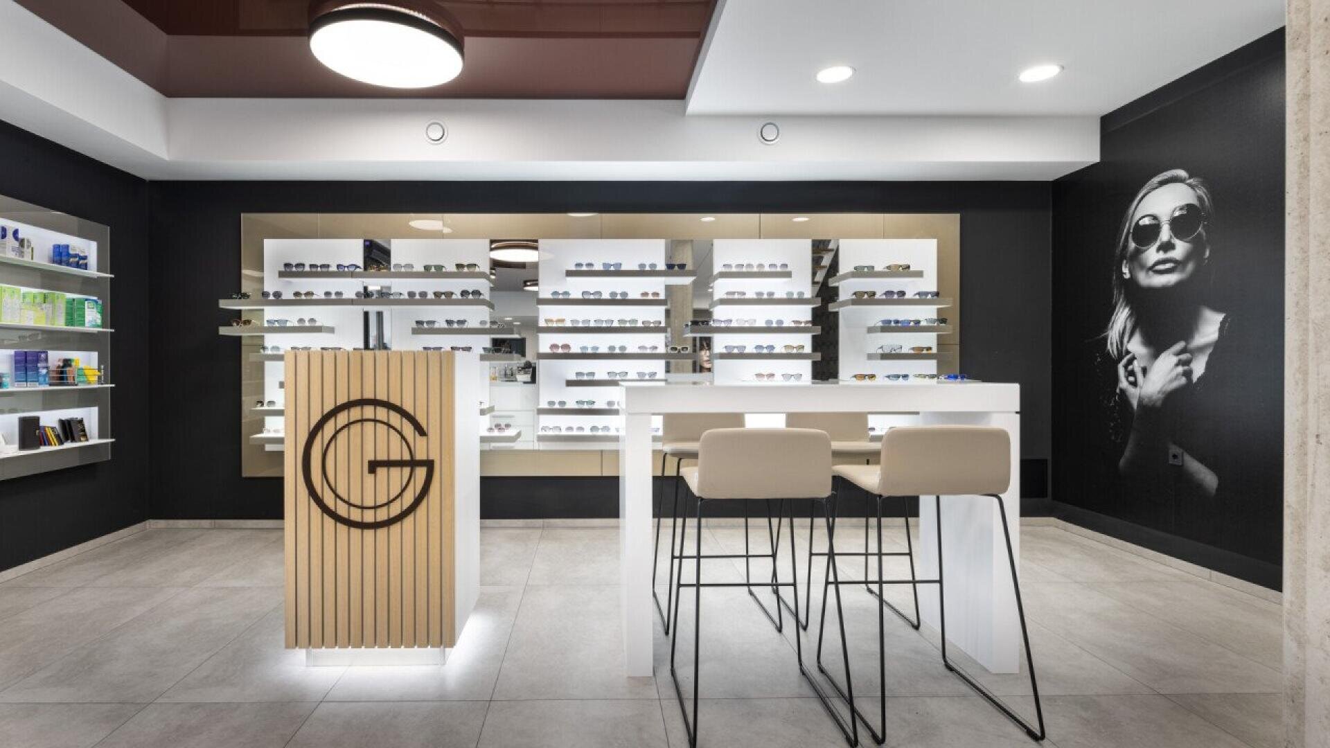 Modern eyewear store interior with display shelves of eyeglasses, a wooden counter with a logo, two high chairs at a white table, and a large black-and-white photo of a woman wearing sunglasses on the wall.