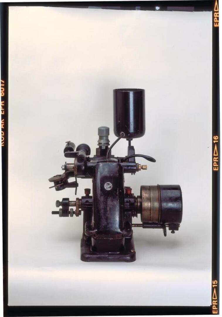 Ein alter schwarzer mechanischer Motor mit verschiedenen Rohren, Ventilen und einem zylindrischen Tank wird vor einem schlichten weißen Hintergrund dargestellt. Die Maschine hat ein metallisches und industrielles Aussehen.