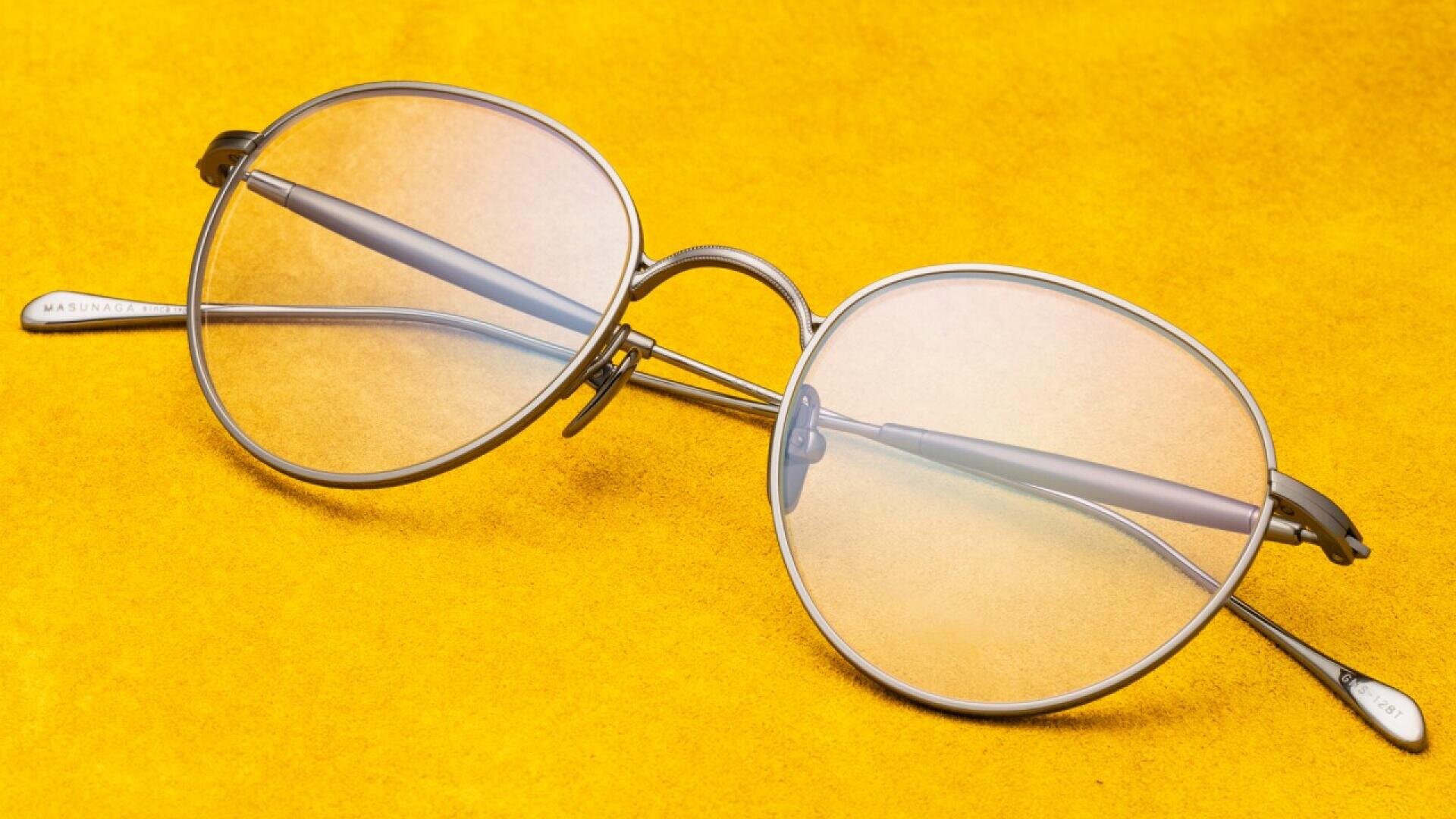 Round, metal-framed eyeglasses with clear lenses rest on a bright yellow surface. The glasses have thin arms and a subtle reflection is visible on the lenses.