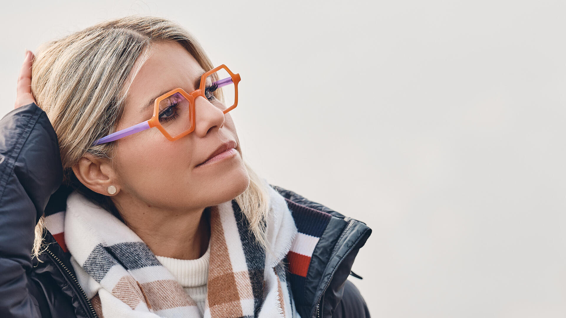Eine Frau mit blondem Haar trägt eine geometrische orange-violette Brille, einen karierten Schal und eine dunkle Jacke. Sie blickt nachdenklich zur Seite, während sie sich durch die Haare fährt, vor einem neutralen, unscharfen Hintergrund.