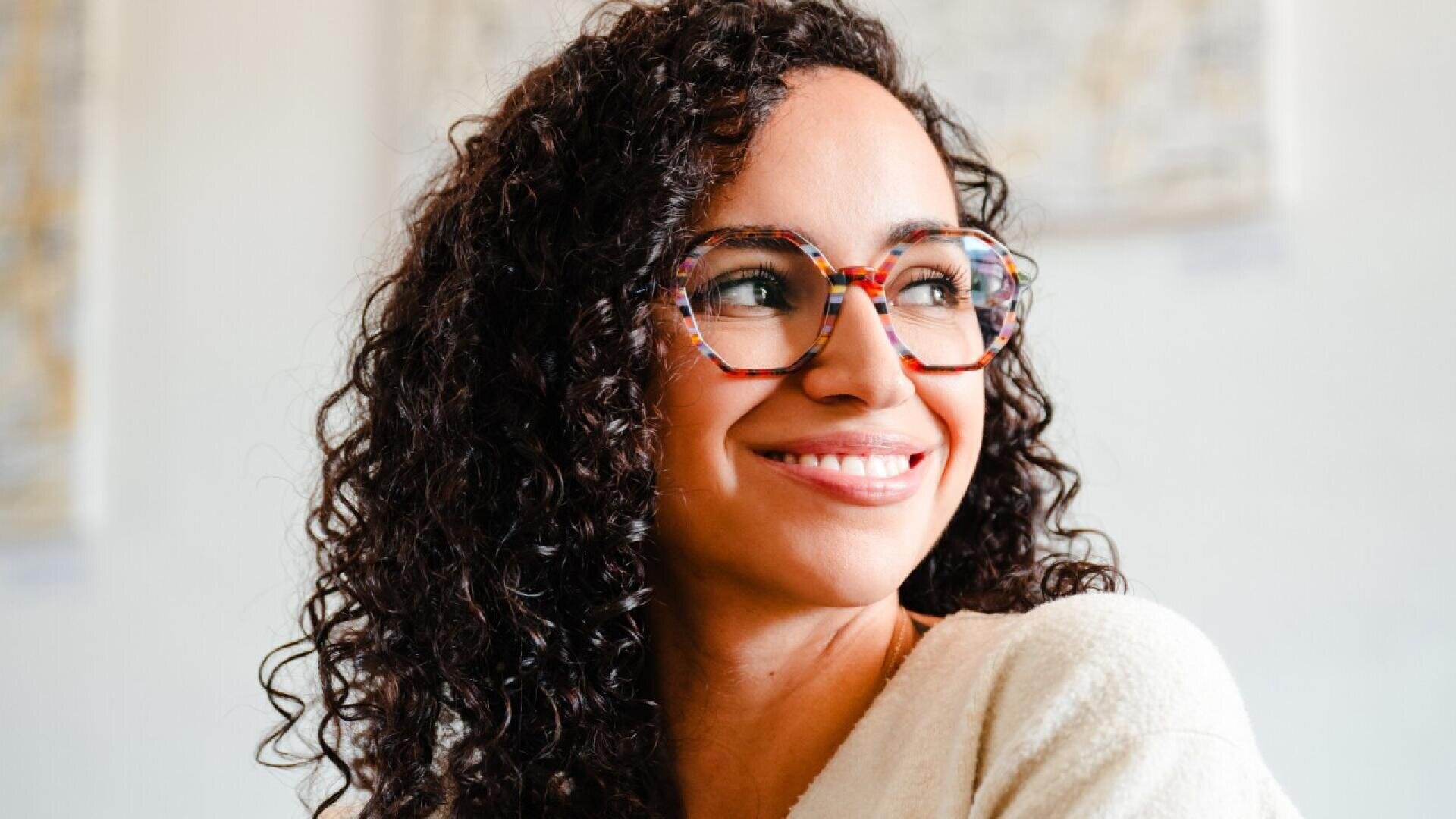 A woman with curly brown hair and patterned eyeglasses smiles while looking to the side. She is wearing a light-colored top and is in a softly lit indoor setting.